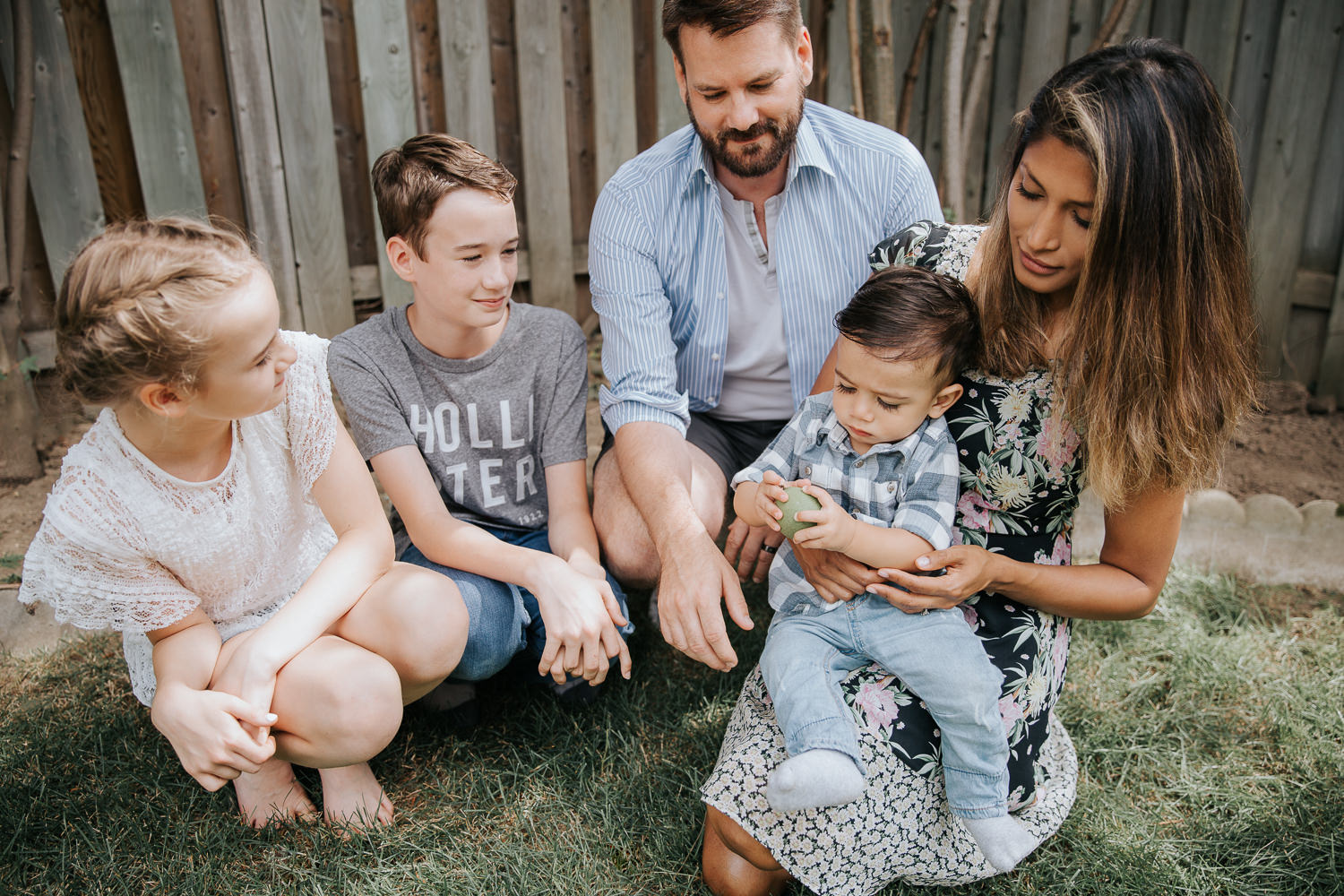 family of 5 in backyard sitting on grass, dad, brother and sister smiling at one year old baby boy sitting in mom' lap holding apple - Markham Lifestyle Photos