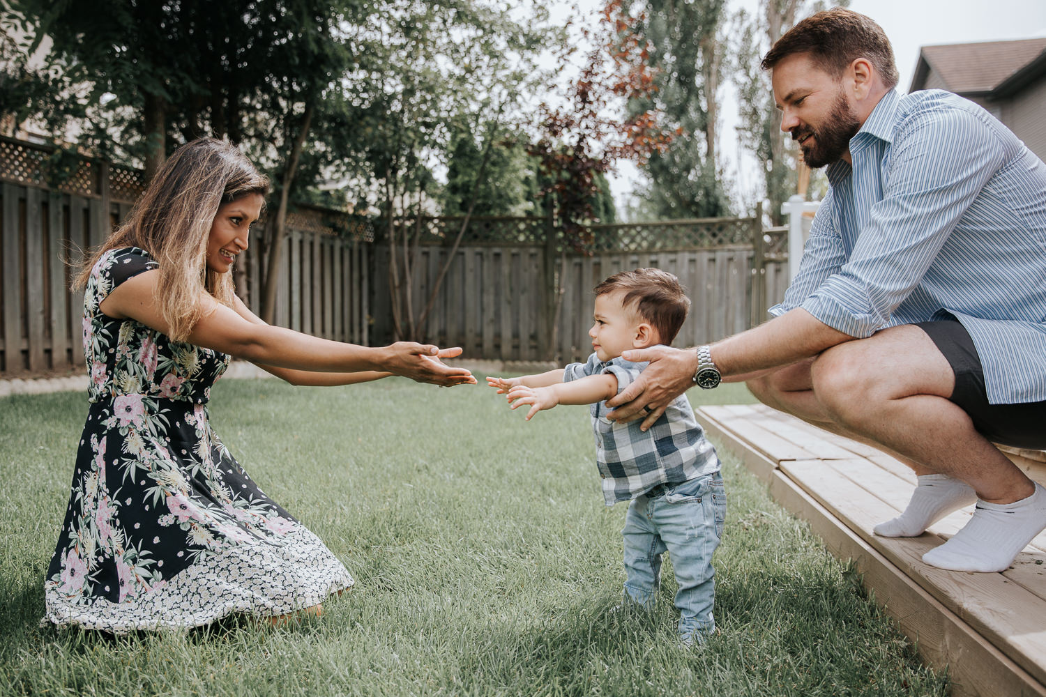 1 year old baby boy with dark hair in white and blue plaid shirt and jeans standing in backyard, supported by dad as mom and son reach out arms to one another, smiling - York Region Lifestyle Photos