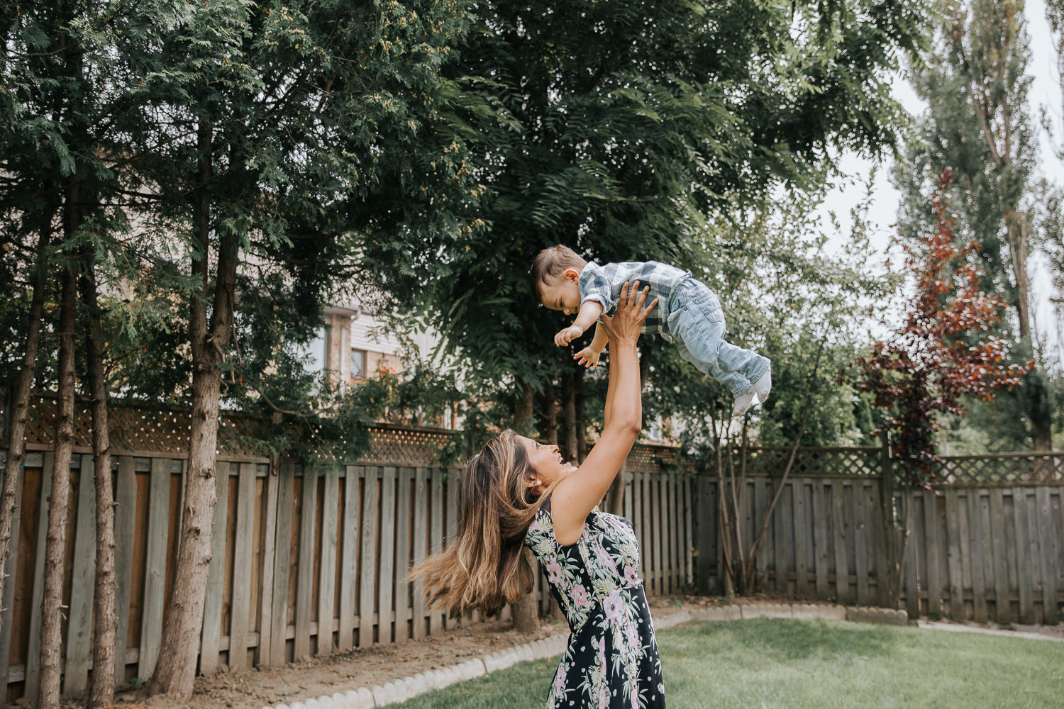 1 year old baby boy with dark hair in white and blue plaid shirt and jeans being thrown in air in backyard by mom - Newmarket Lifestyle Photography