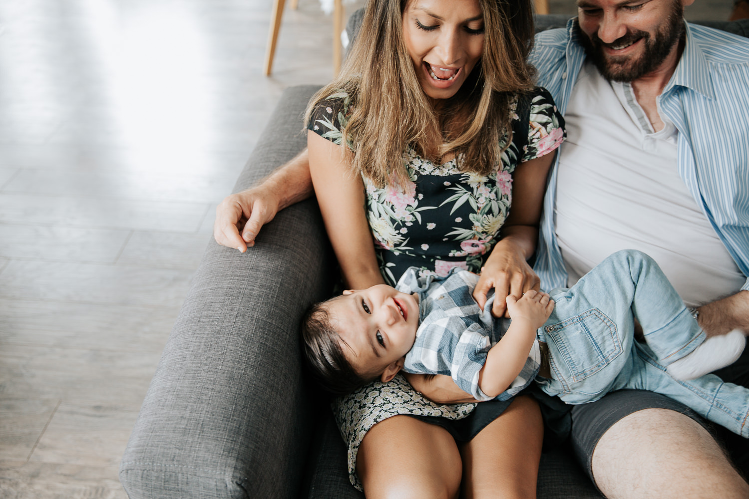 mother and father sitting on couch playing with 1 year old baby boy with dark hair sitting in mom's lap, mother tickling son who is smiling at camera - GTA Lifestyle Photography