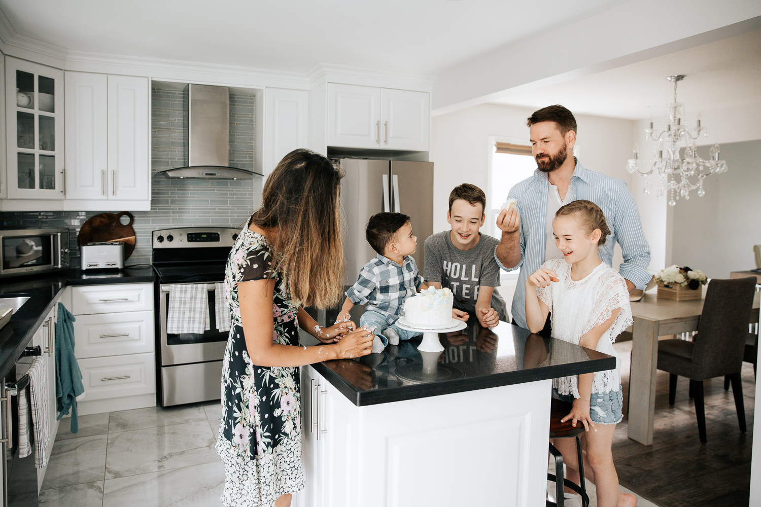 family of 5 standing around island in kitchen watching 1 year old baby boy start cake smash, dad eating piece of cake - Newmarket In-Home Photos