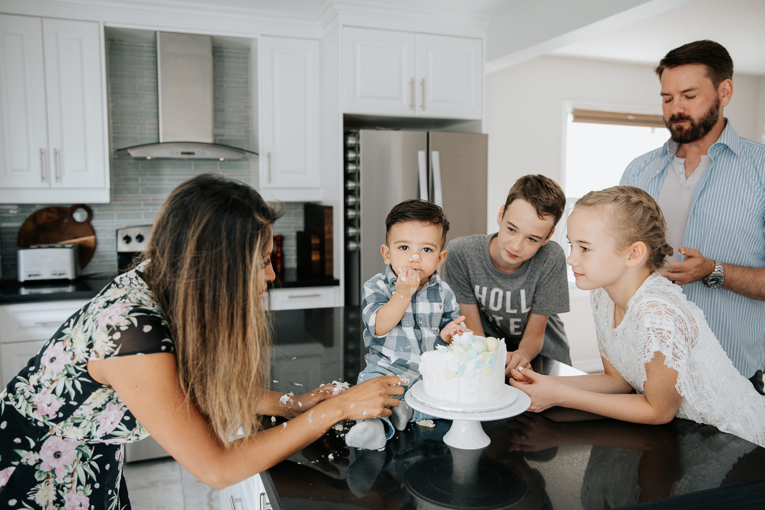family of 5 standing around island in kitchen watching 1 year old baby boy doing first birthday cake smash - Markham In-Home Photos