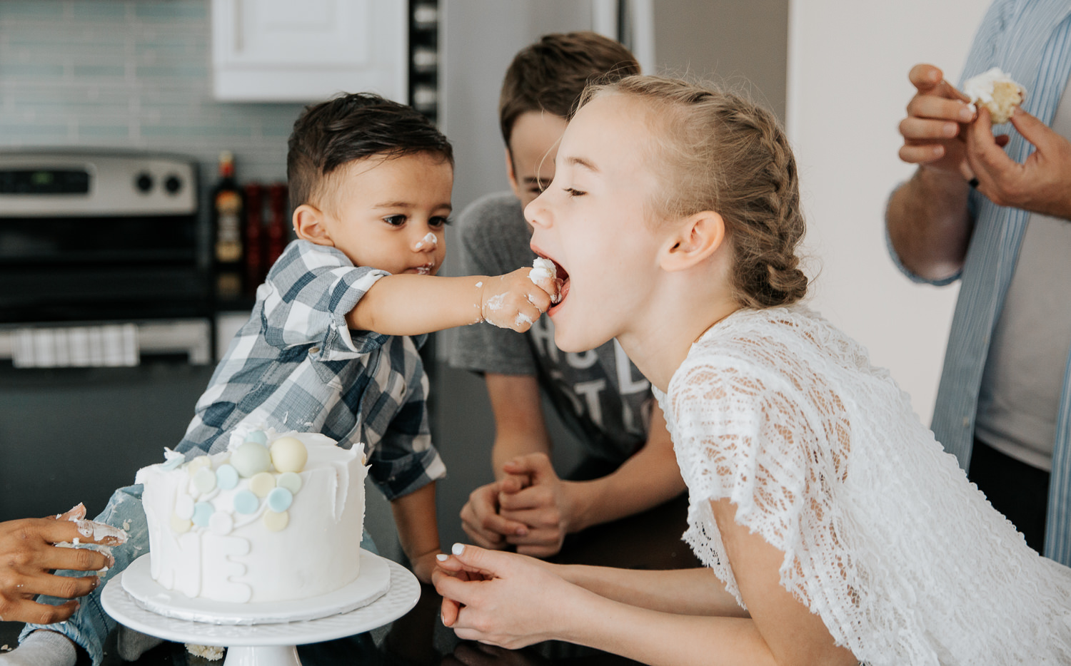 1 year old baby boy with dark hair sitting on kitchen counter doing first birthday cake smash, feeding cake to 11 year old big sister - Stouffville In-Home Photos