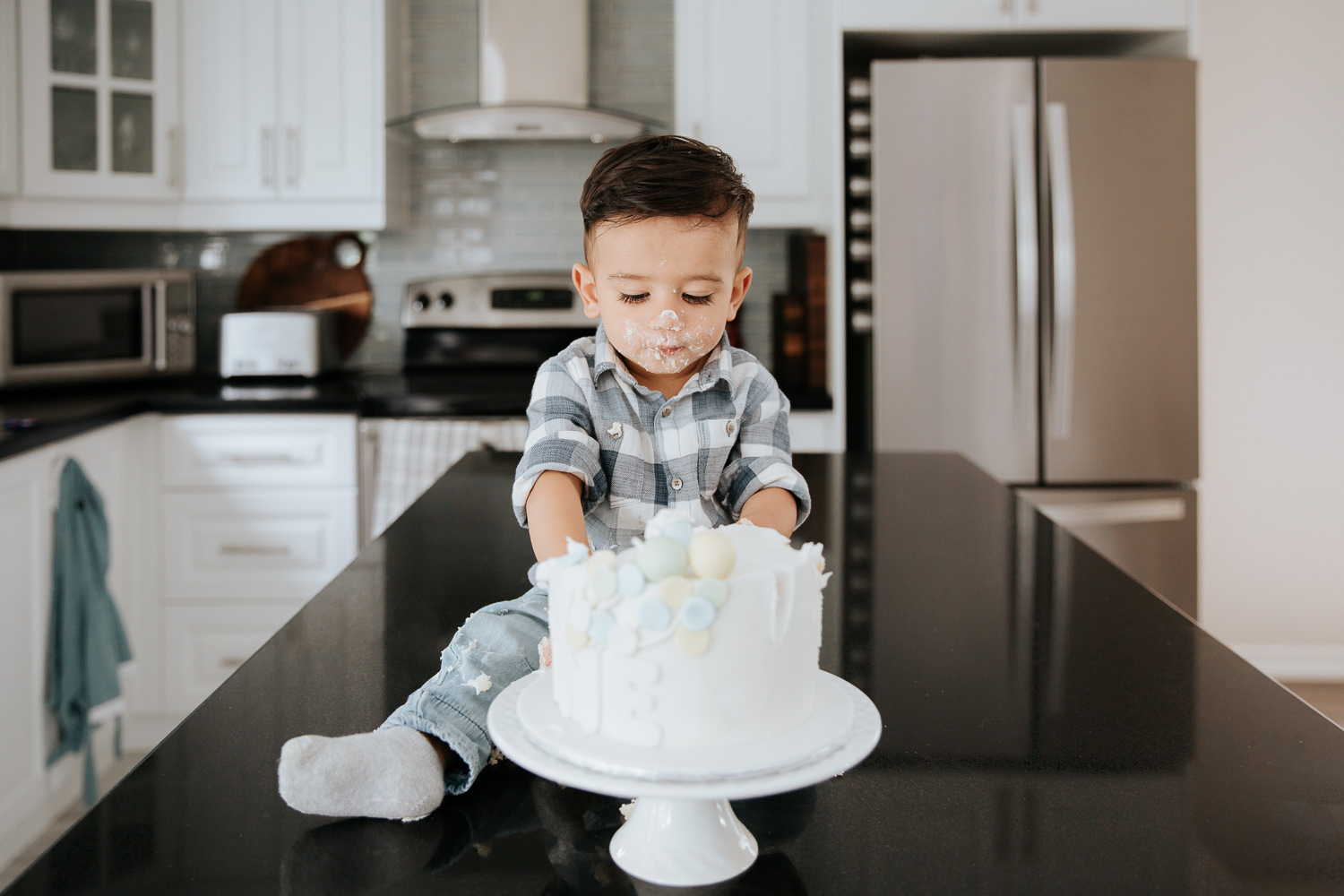 1 year old baby boy with dark hair sitting on kitchen counter eating cake for first birthday - Barrie In-Home Photos
