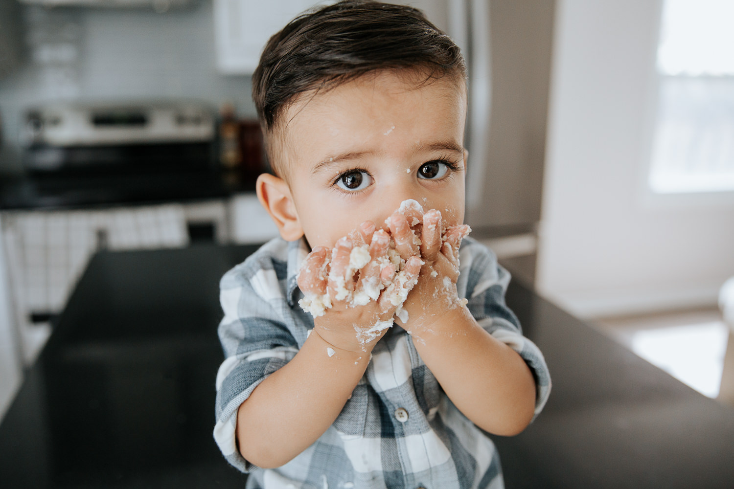 1 year old baby boy with dark brown hair and eyes in blue and white plaid shirt looking at camera with hands covered in cake up at mouth - GTA In-Home Photos