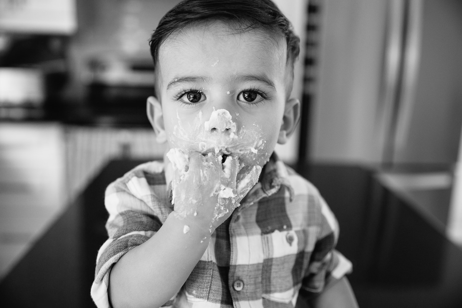 York Region In-Home Photos1 year old baby boy with dark brown hair and eyes in blue and white plaid shirt looking at camera with hand covered in cake up in mouth - 