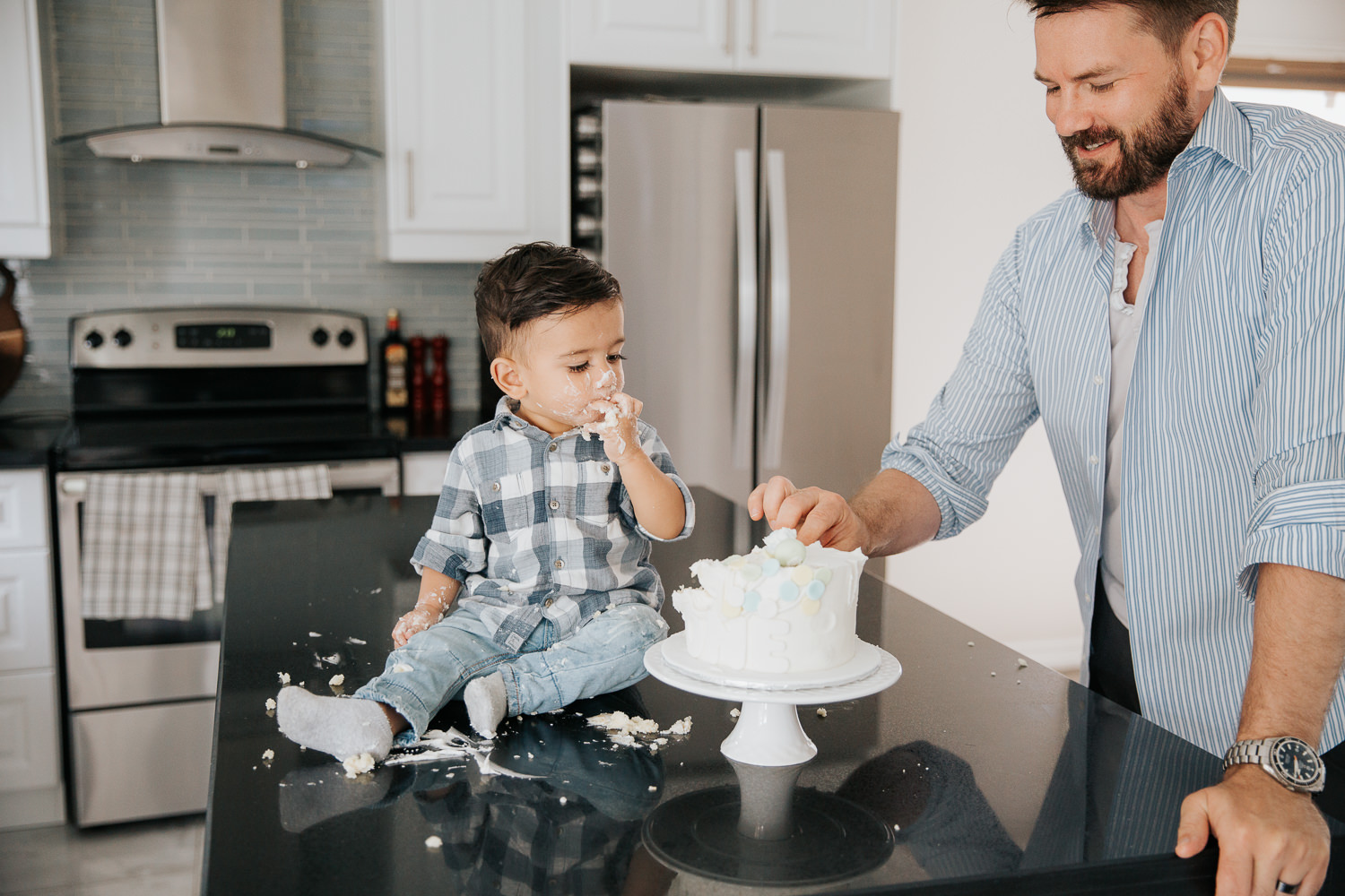 1 year old baby boy with dark brown hair and eyes in blue and white plaid shirt putting cake in his mouth and watching dad take piece - Newmarket In-Home Photography