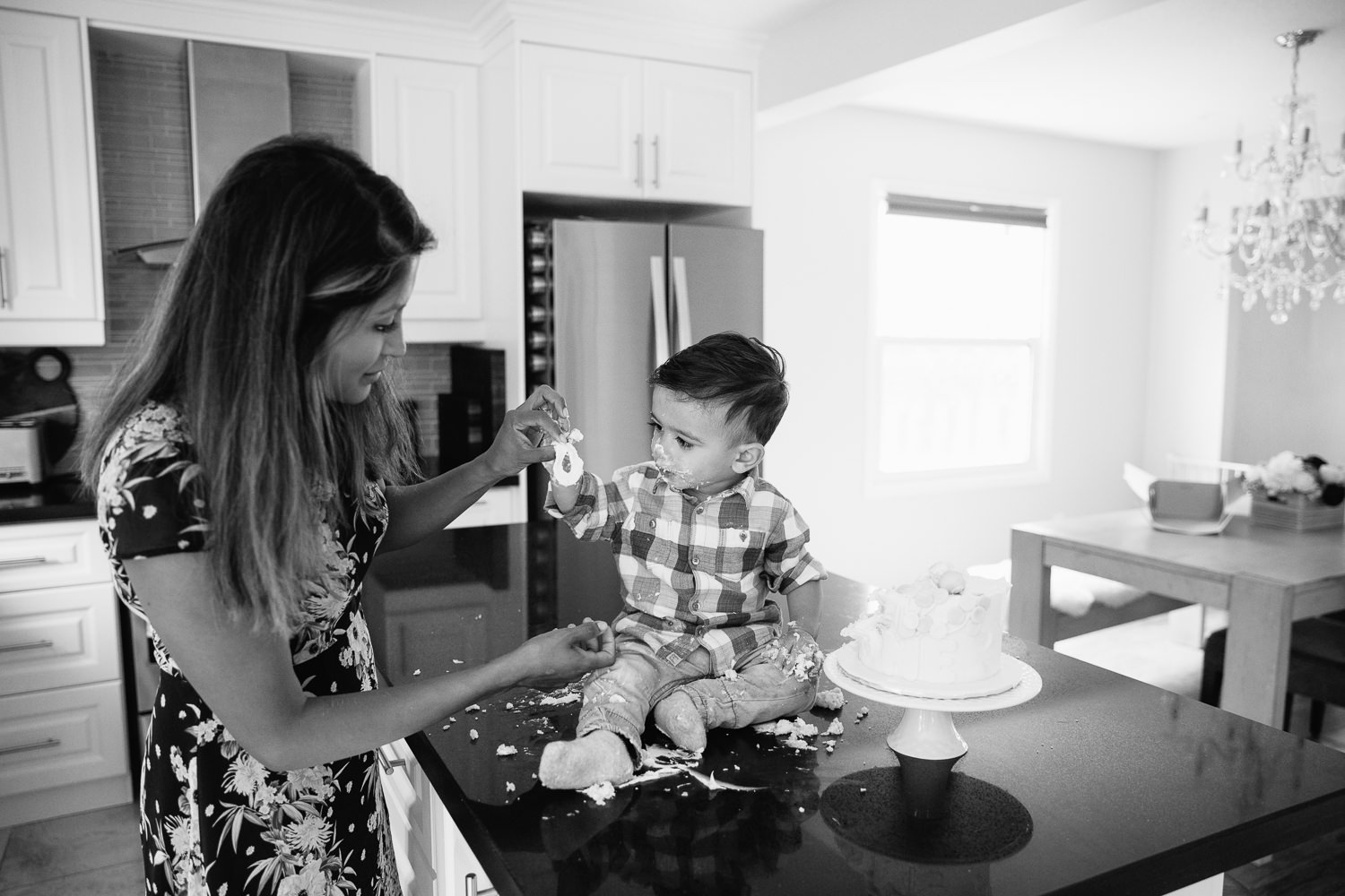 1 year old baby boy with dark brown hair and eyes in blue and white plaid shirt sitting on kitchen counter doing cake smash and playing with icing with mom - Stouffville In-Home Photography