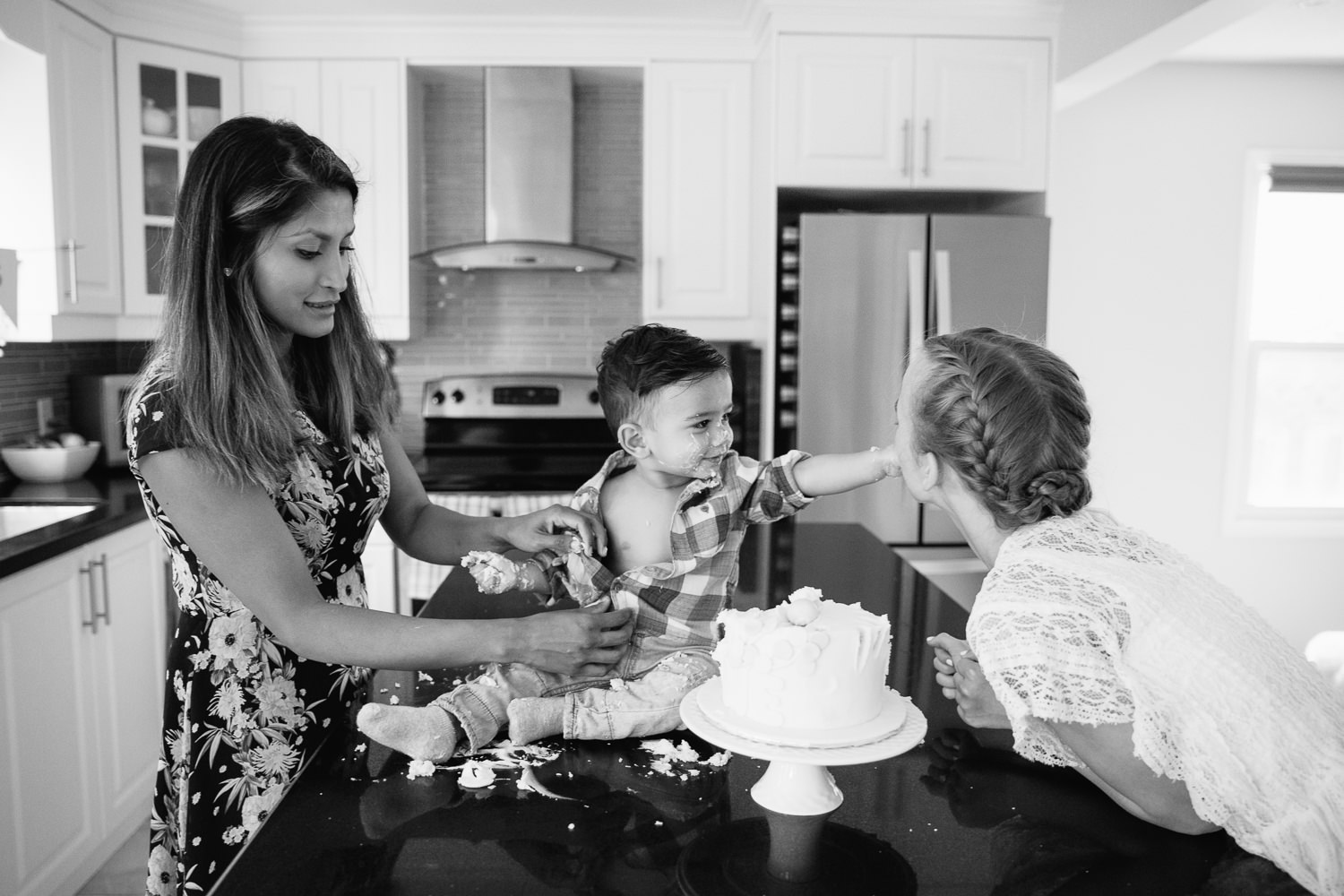 1 year old baby boy with dark brown hair and eyes in plaid shirt sitting on kitchen counter doing cake smash as mom takes off his shirt, son is feeding big sister - Markham In-Home Photography