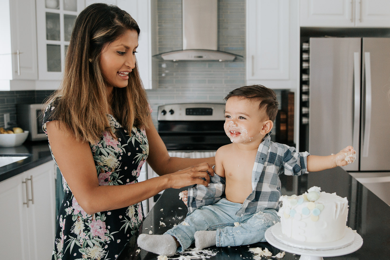 1 year old baby boy with dark brown hair and eyes in blue and white plaid shirt sitting on kitchen counter doing cake smash as mom tries to take off his shirt - Barrie In-Home Photography