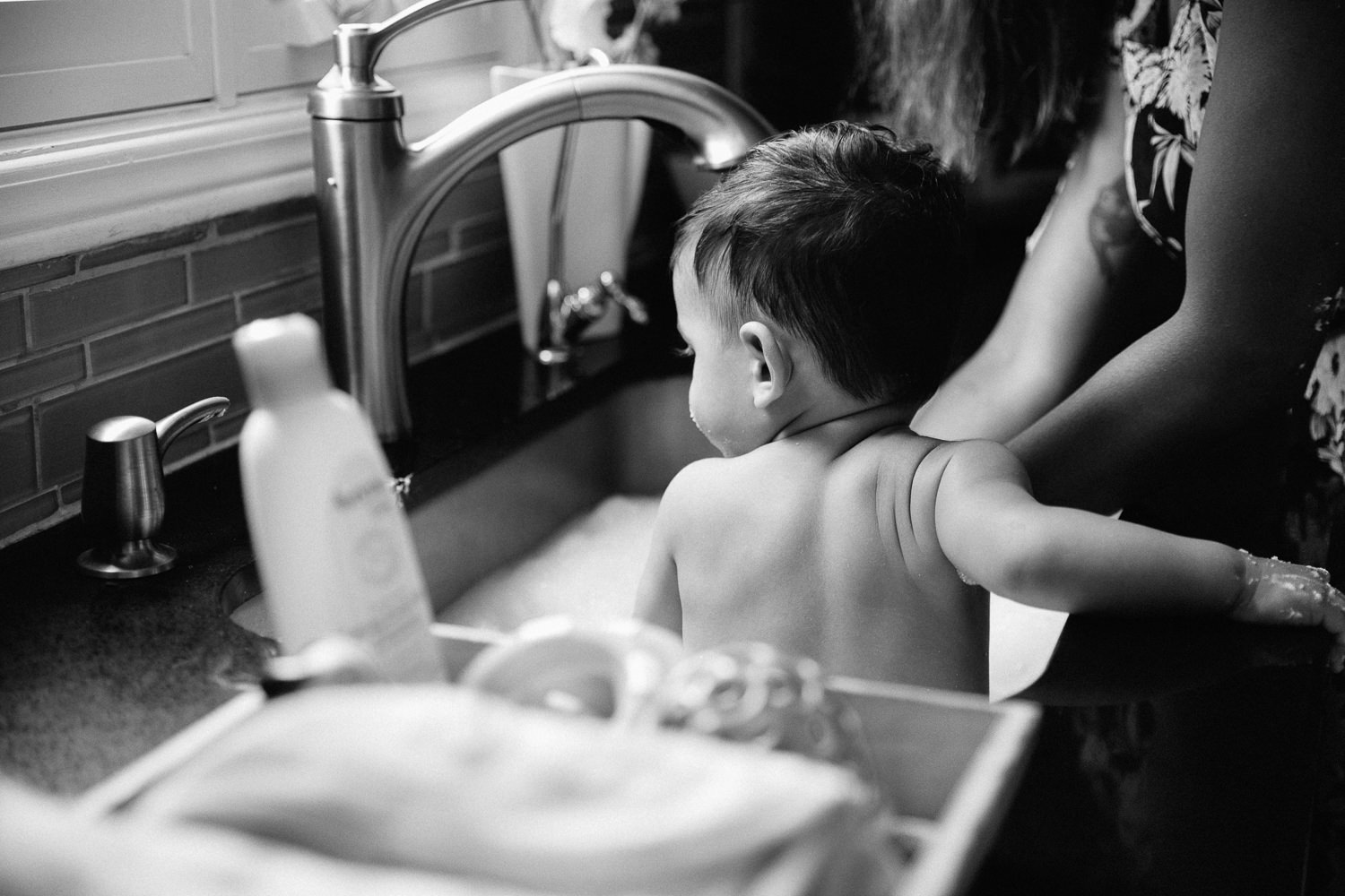 1 year old baby boy with dark hair sitting in kitchen sink as mom gives him a bath, back to camera - York Region In-Home Photography