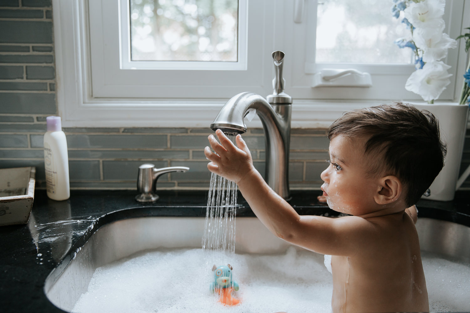 1 year old baby boy with dark hair and eyes sitting in kitchen sink for a bath, holding hand under water running from faucet - Markham Lifestyle Photography
