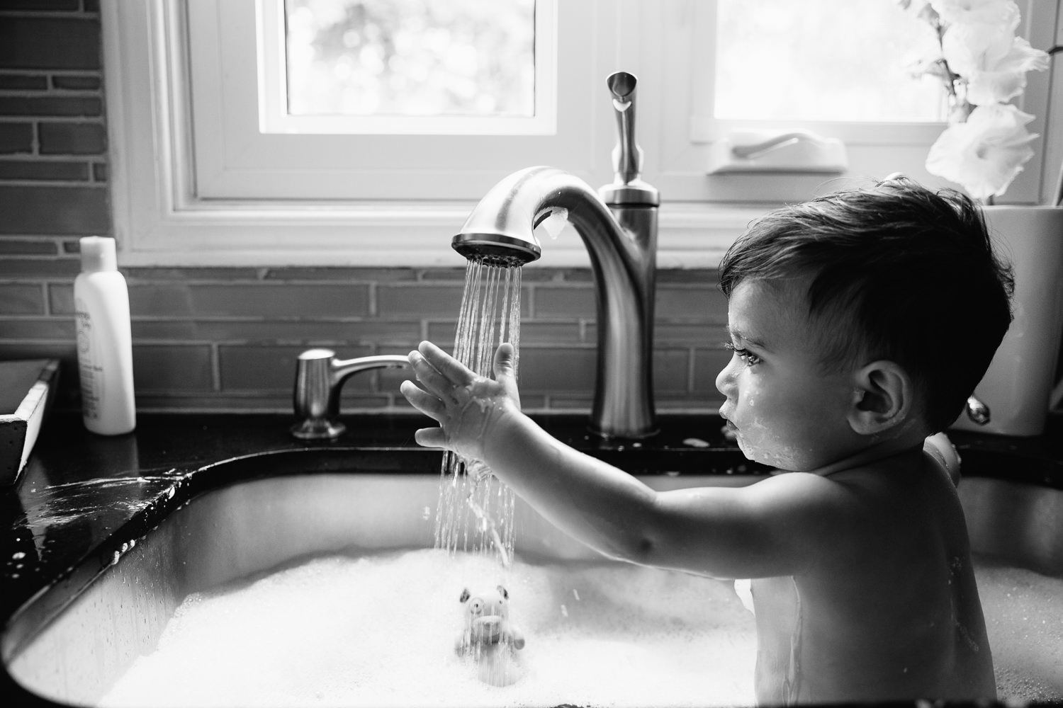 1 year old baby boy with dark hair and eyes sitting in kitchen sink for a bath, holding hand under water running from faucet - Barrie Lifestyle Photography