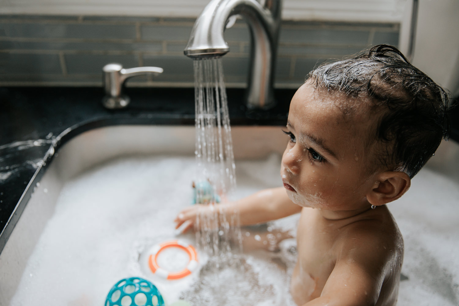 1 year old baby boy with dark hair and eyes sitting in kitchen sink for a bath, water running sink full of bubbles - GTA Lifestyle Photography