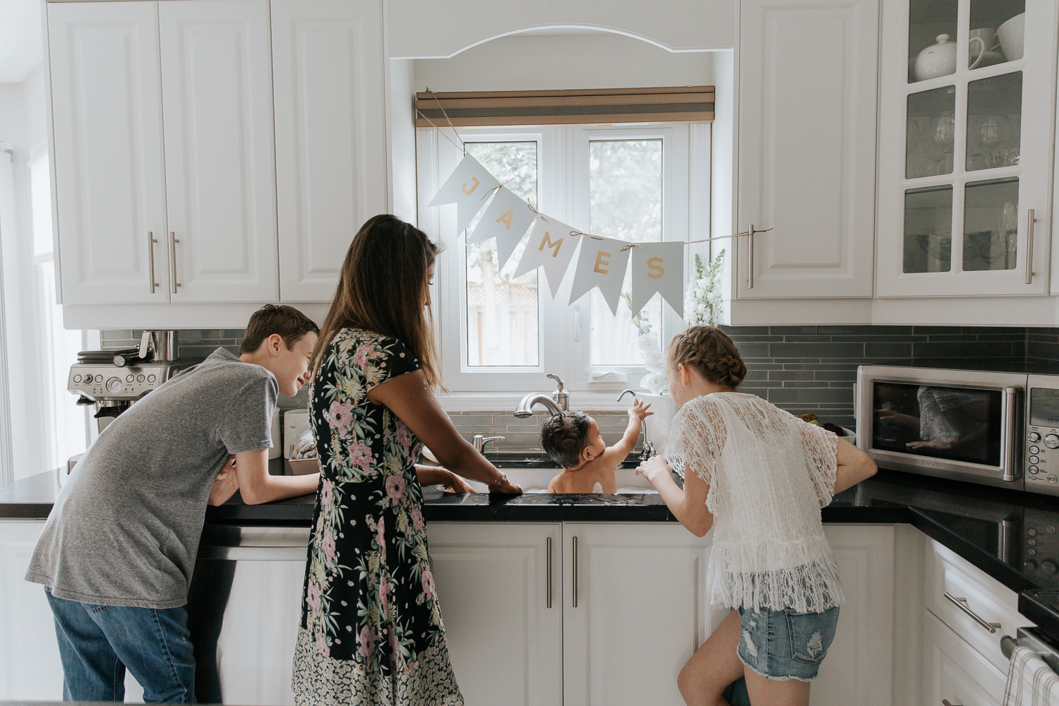 mom, 11 year old girl and 13 year old boy standing at kitchen sink giving 1 year old baby brother a bath - York Region Lifestyle Photography