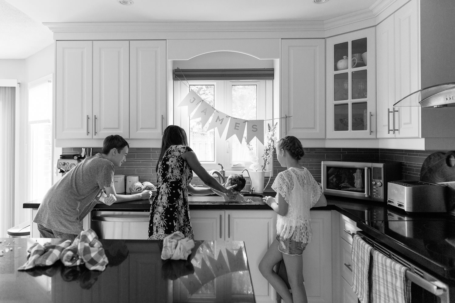 mom, 11 year old girl and 13 year old boy standing at kitchen sink giving 1 year old baby brother a bath - Newmarket Lifestyle Photos