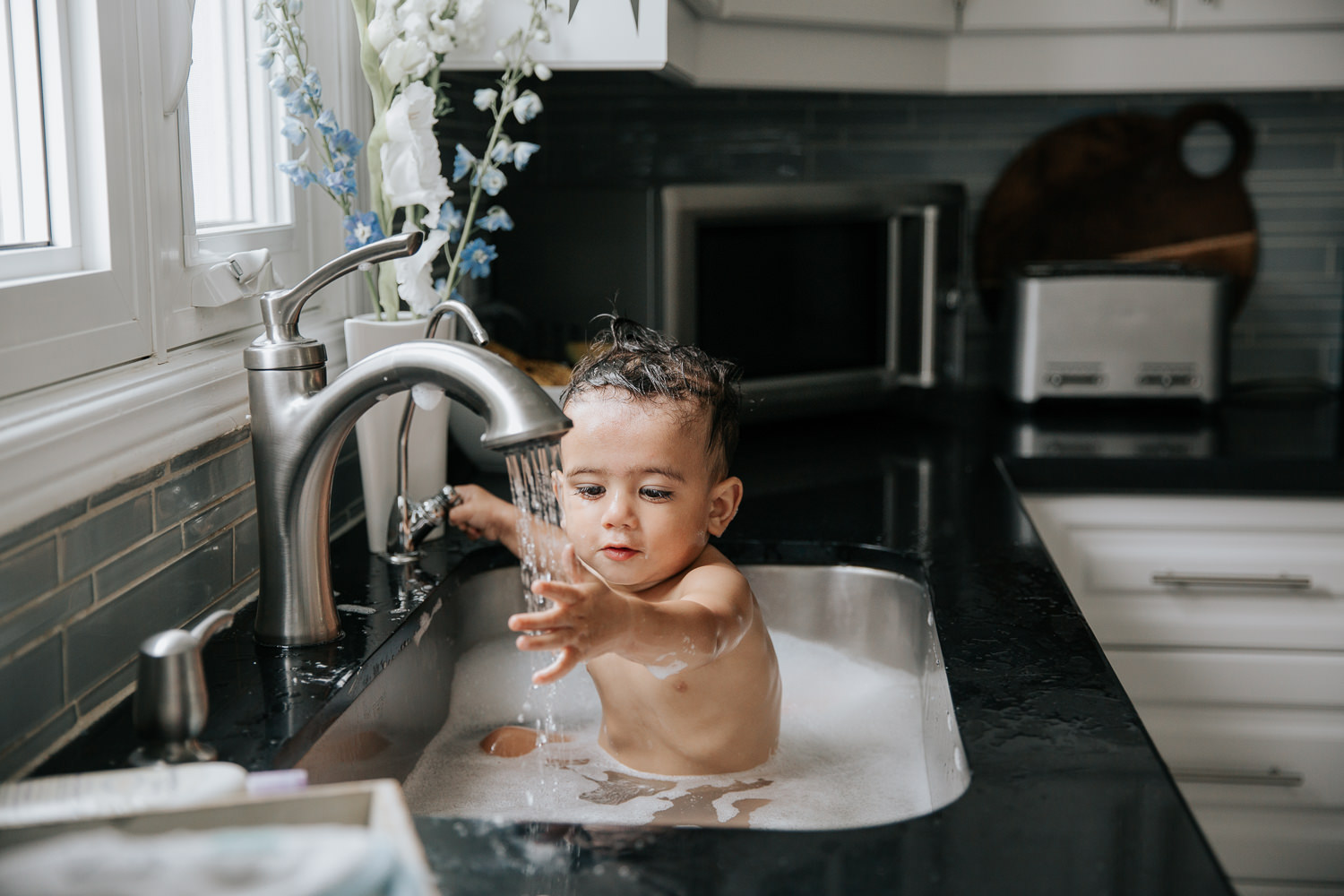 1 year old baby boy with dark hair and eyes sitting in kitchen sink for a bath, holding hand under water running from faucet - Markham Lifestyle Photos