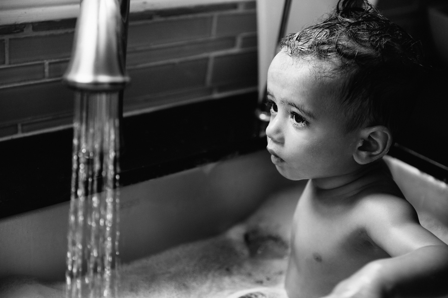 1 year old baby boy with dark hair and eyes sitting in kitchen sink for a bath, water running from faucet - Barrie Lifestyle Photos