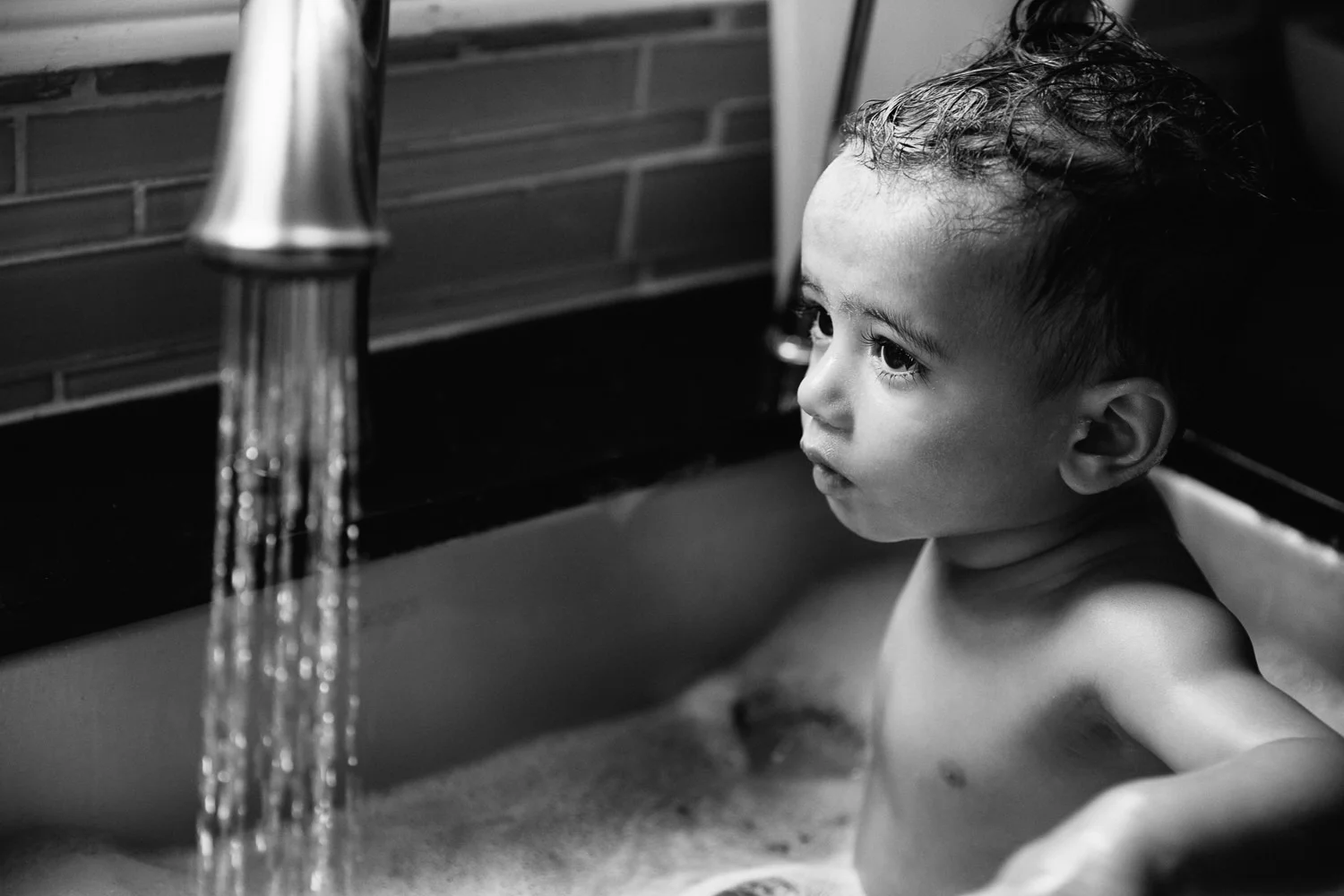 1 year old baby boy with dark hair and eyes sitting in kitchen sink for a bath, water running from faucet - Barrie Lifestyle Photos