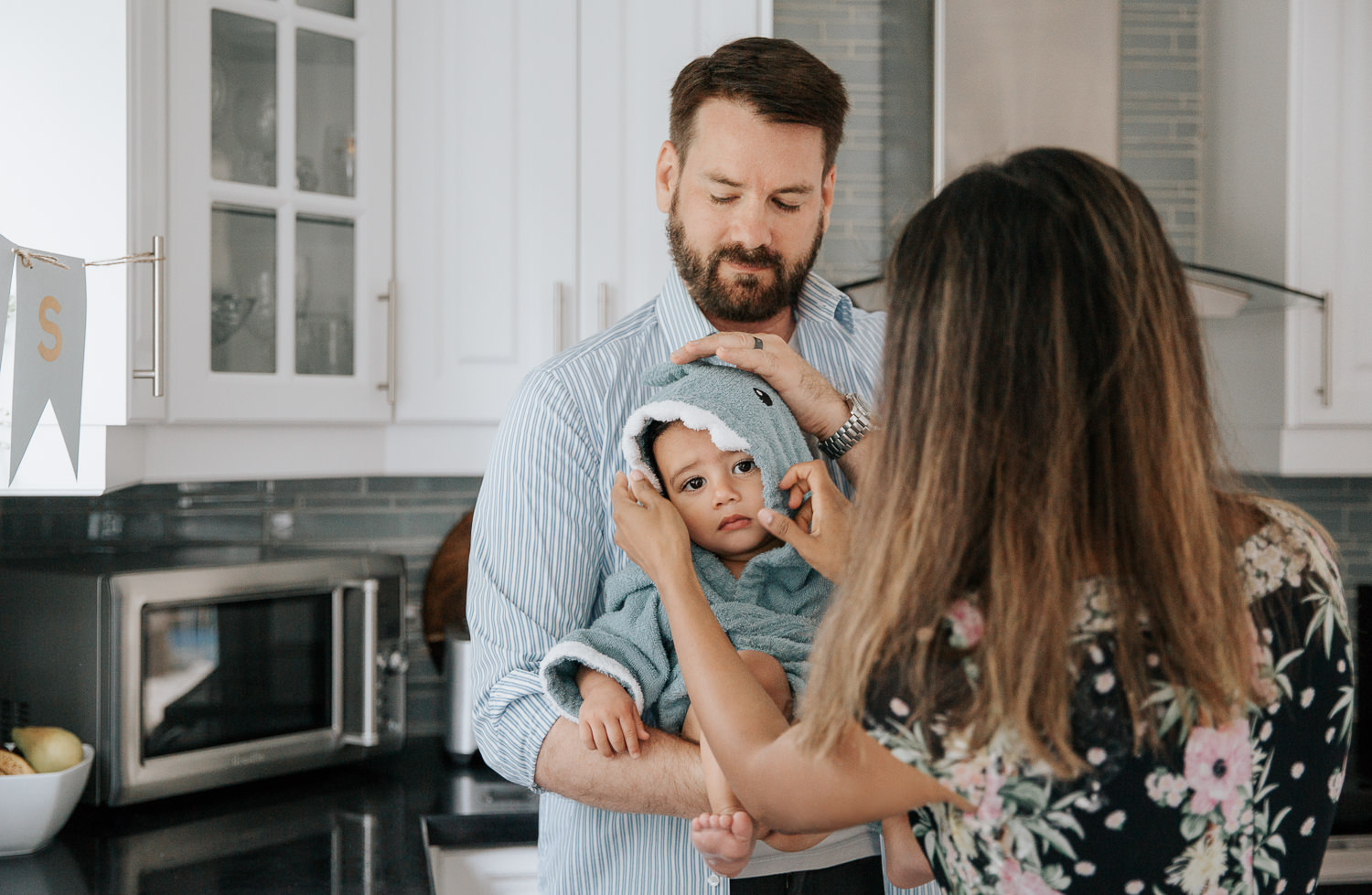 dad standing in kitchen holding 1 year old baby boy fresh out of bath, mom wrapping son in blue shark hooded towel - GTA Lifestyle Photos