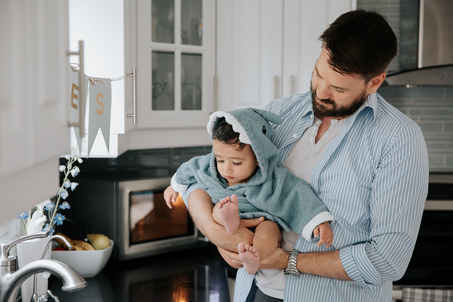 dad standing in kitchen holding 1 year old baby boy fresh out of bath, son wrapped in blue shark hooded towel - York Region Lifestyle Photos