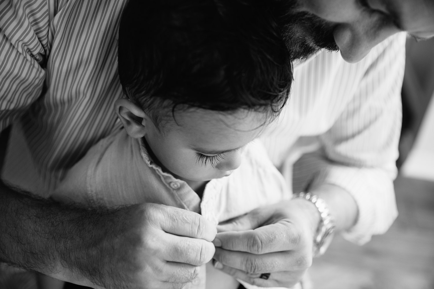 1 year old baby boy with dark hair and long eyelashes sitting in dad's lap as father button's son's shirt - Barrie In-Home Photos