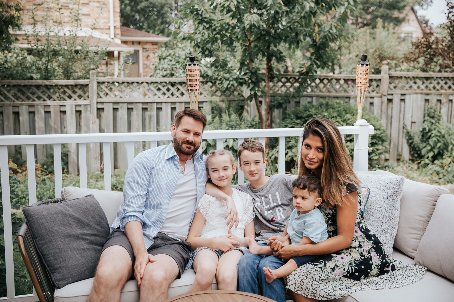 family of 5 sitting on couch on backyard deck, 11 year old girl, 13 year old boy and 1 year old baby sitting between parents, smiling at camera - Markham Lifestyle Photography