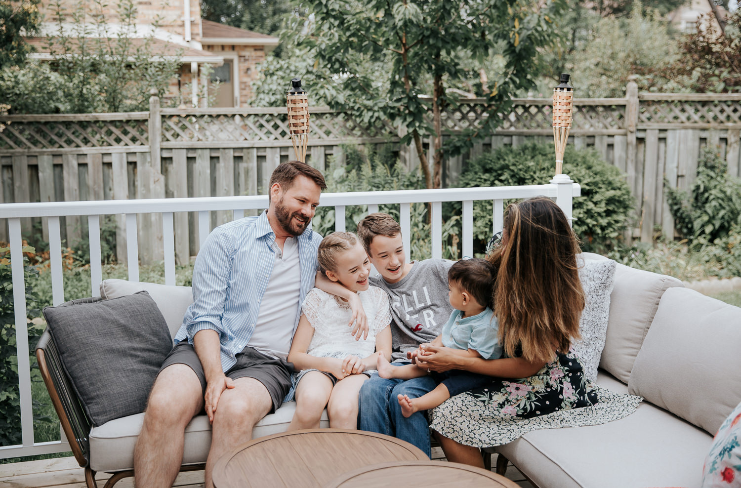 family of 5 sitting on couch on backyard deck, 11 year old girl, 13 year old boy and 1 year old baby sitting between parents, everyone looking at each other laughing - Barrie Lifestyle Photography