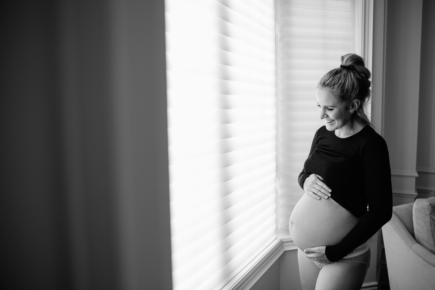 pregnant woman with blonde hair in braid wearing black top, baby bump exposed, holding belly and smiling standing next to window - Stouffville Lifestyle Photography