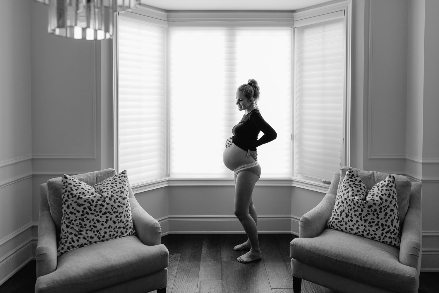 pregnant woman with blonde hair in braid wearing black top and grey underwear, baby bump exposed, holding belly and smiling standing in front of window - GTA Lifestyle Photography