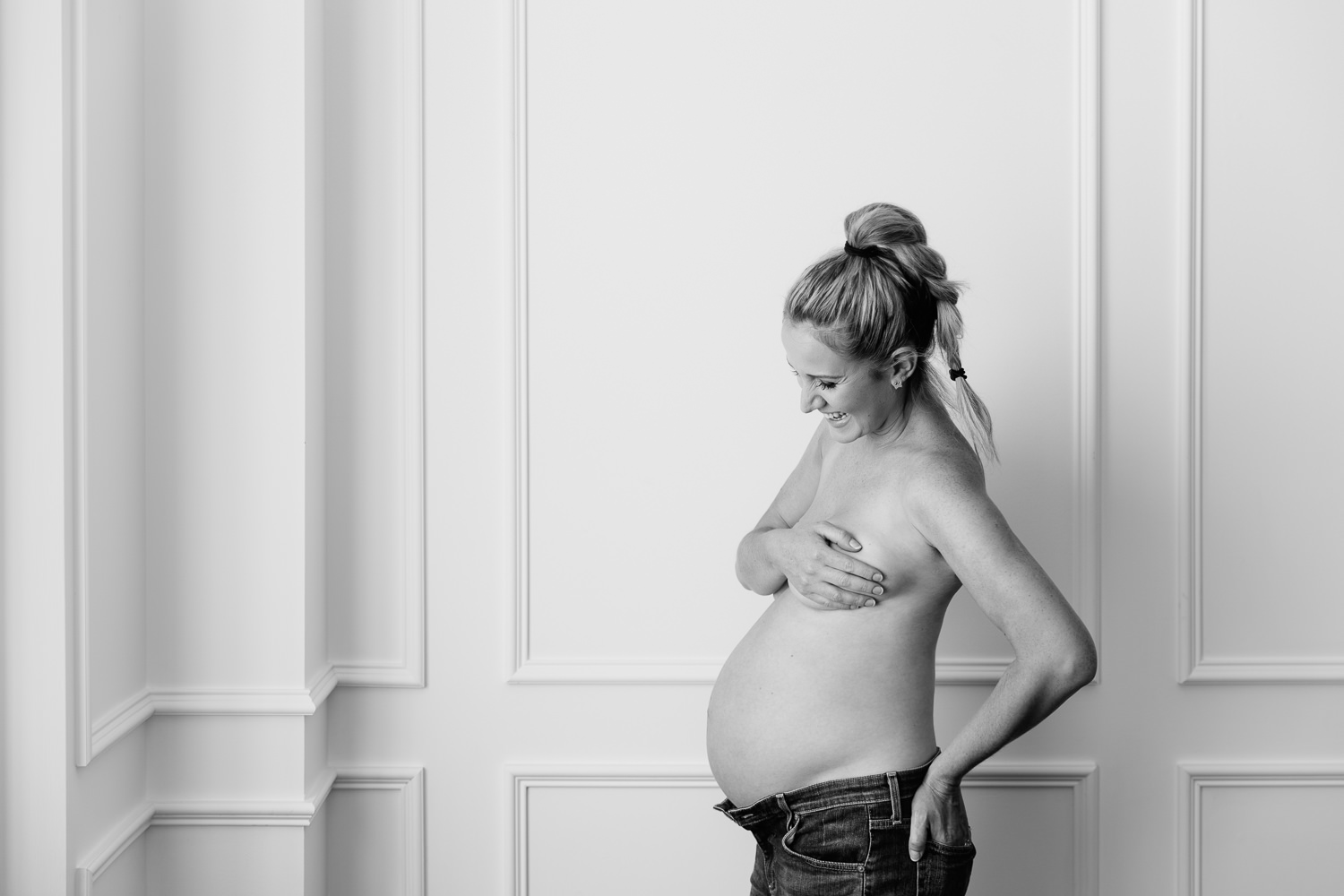 pregnant woman with blonde hair in braid wearing jeans, standing in front of while wall laughing, baby bump exposed - Markham In-Home Photography