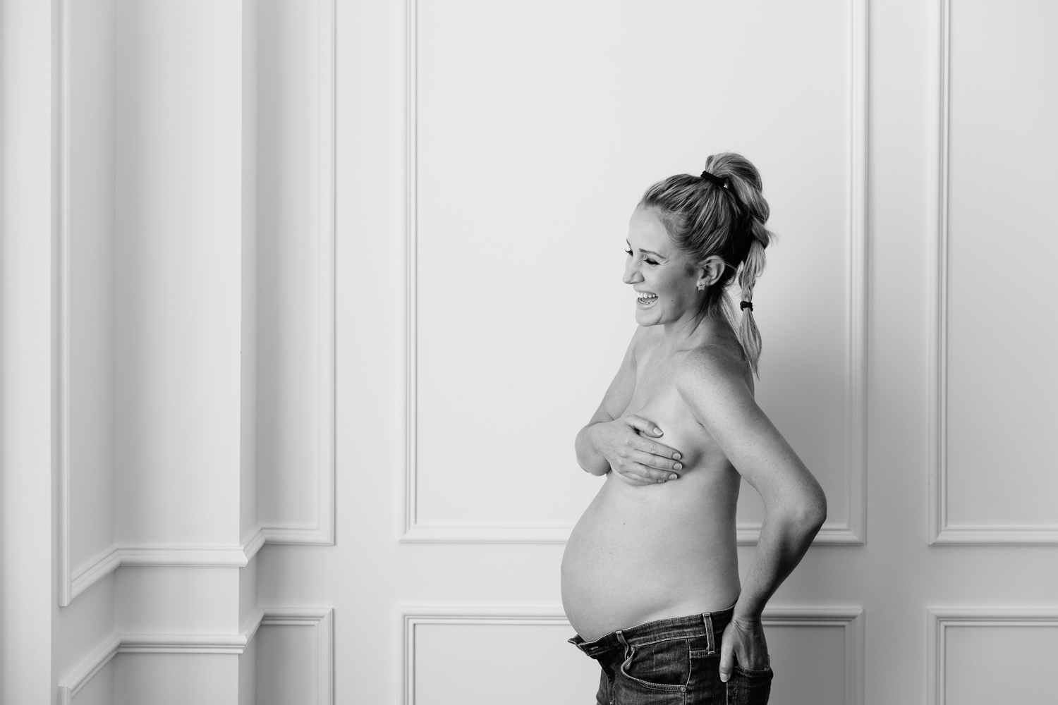 pregnant woman with blonde hair in braid wearing jeans, standing in front of while wall laughing, baby bump exposed - Stouffville In-Home Photography