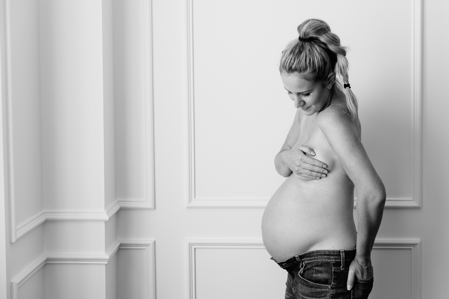 pregnant woman with blonde hair in braid wearing jeans, standing in front of while wall laughing, baby bump exposed - Barrie In-Home Photography