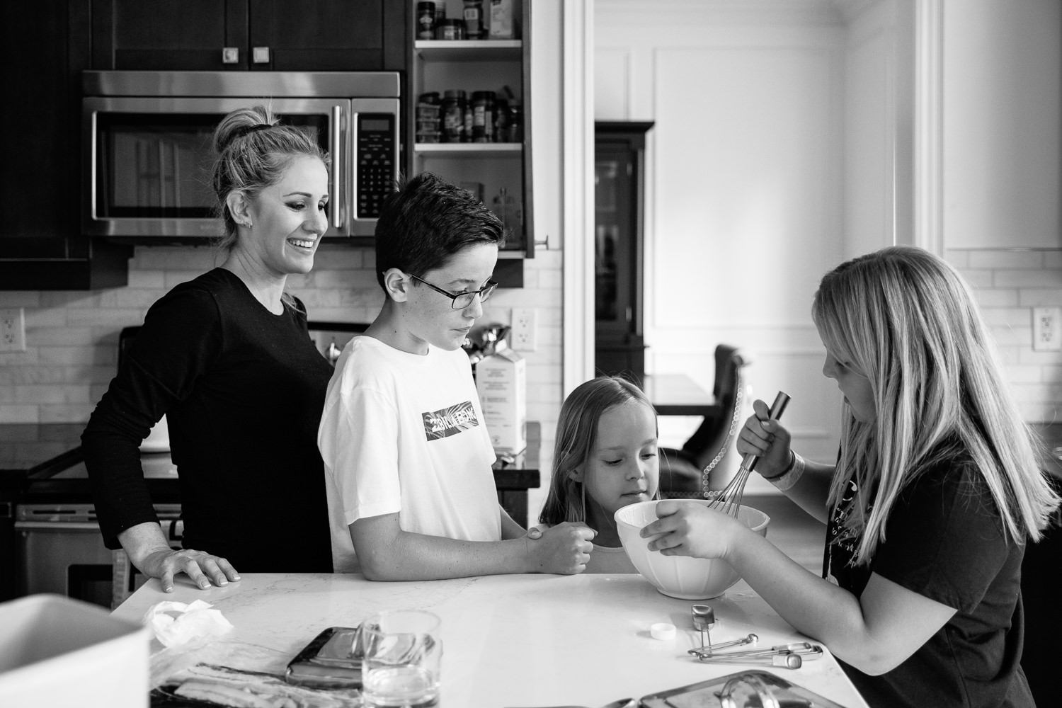3 children standing at kitchen island mixing pancake batter for family brunch, 8 and 10 year old girls, mom standing behind 13 year old boy watching - Markham Lifestyle Photography