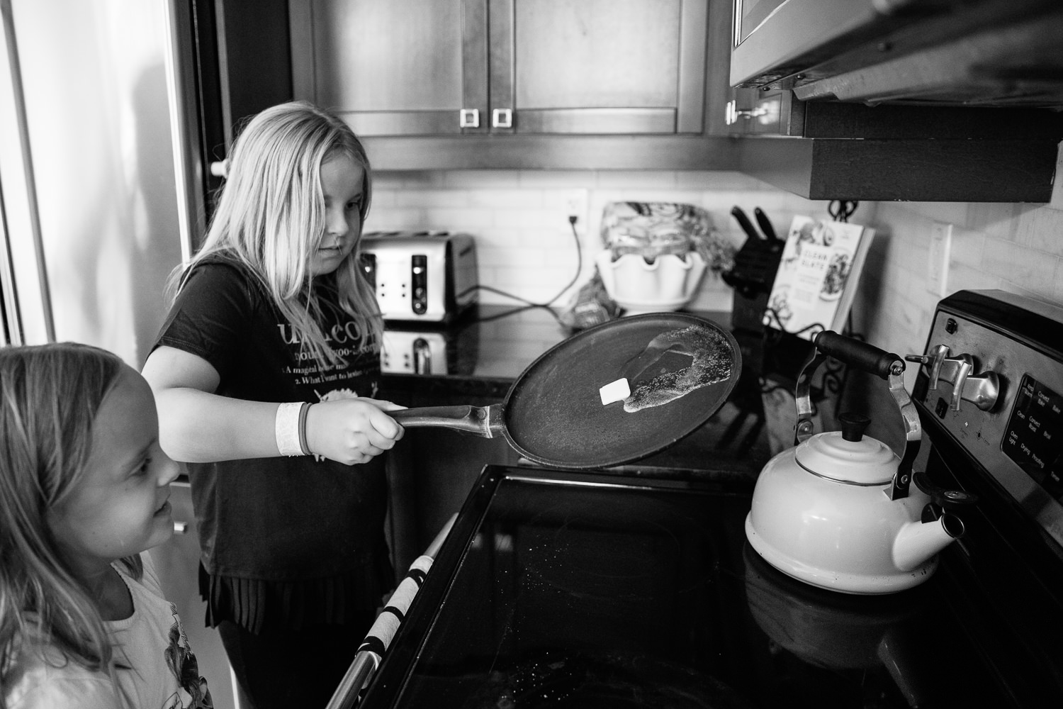 8 and 10 year old girls standing at stove, older sister melting butter across hot pan - Barrie Lifestyle Photography