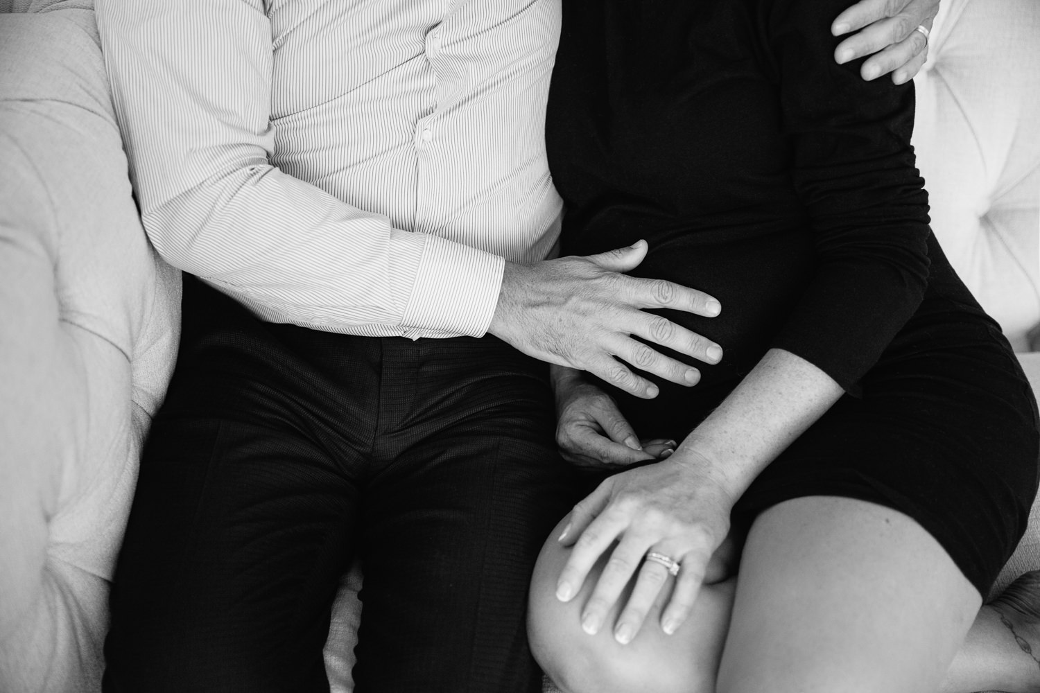 couple sitting together on couch, husband's hand on pregnant wife's belly, woman wearing black dress - Markham In-Home Photography