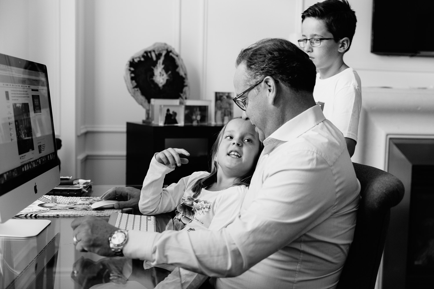 8 year old girl sitting in dad's lap as they sit at his desk, daughter looking up and talking to father, older brother standing behind them - York Region Lifestyle Photography