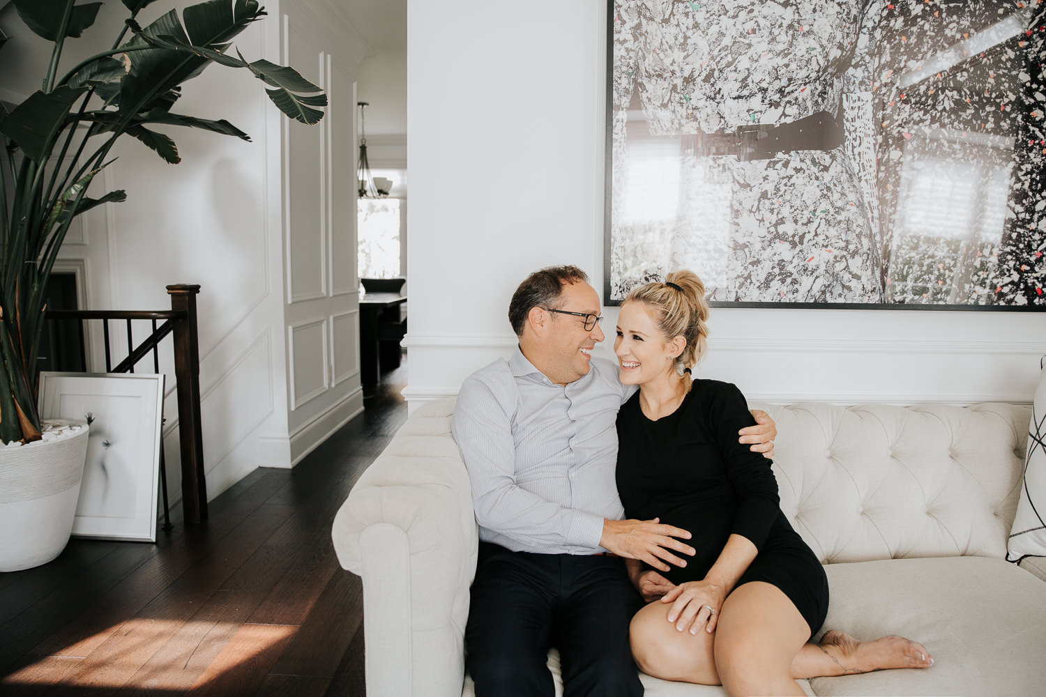 couple sitting together on couch smiling at one another, husband's hand on pregnant wife's belly, woman wearing black dress and blonde hair in braid - Stouffville In-Home Photography