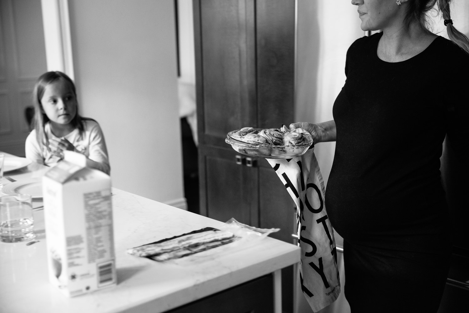 pregnant woman in black dress carrying pie plate of cinnamon buns to kitchen island for family brunch - York Region In-Home Photos