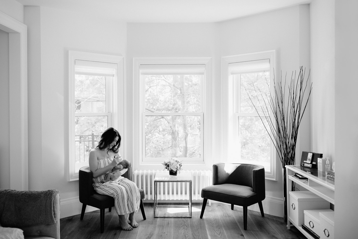 new mom with long dark hair sitting in chair in white, light filled room in front of window bottle feeding 2 week old baby boy in diaper - Newmarket Lifestyle Photography