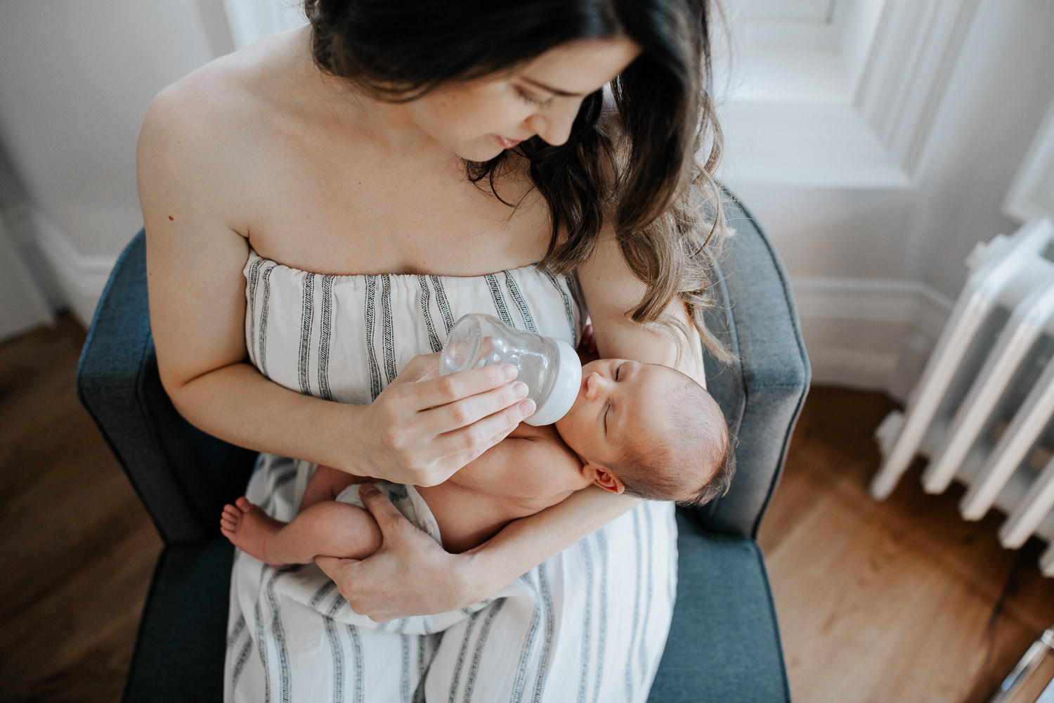 new mom with long dark hair sitting in chair in white, light filled room in front of window bottle feeding 2 week old baby boy in diaper - Stouffville Lifestyle Photography