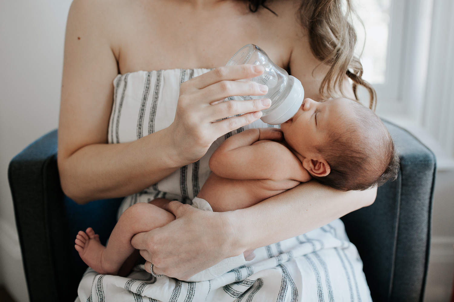 new mom sitting in chair in white, light filled room bottle feeding 2 week old baby boy in diaper - GTA Lifestyle Photography