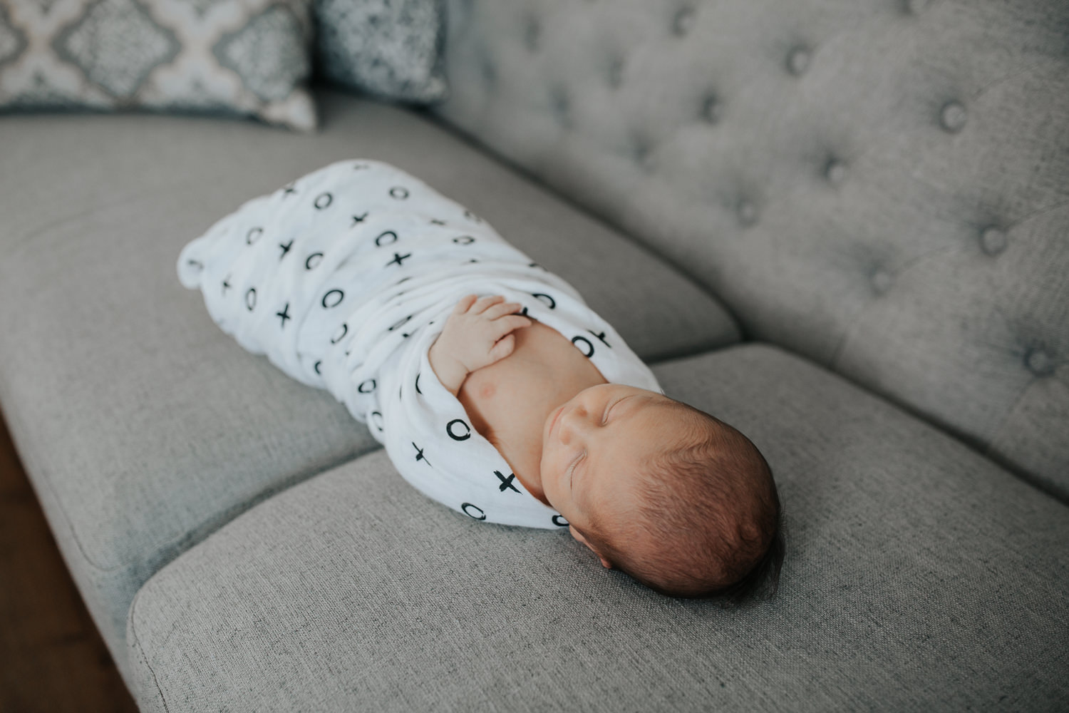 2 week old baby boy with dark hair wrapped in black and white swaddle with arm out, sleeping on grey couch in living room - Barrie In-Home Photography