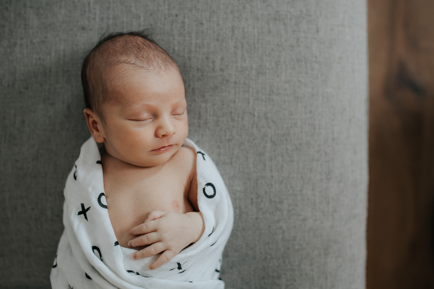 2 week old baby boy with dark hair wrapped in black and white swaddle with arm out, sleeping on grey couch in living room - GTA In-Home Photography