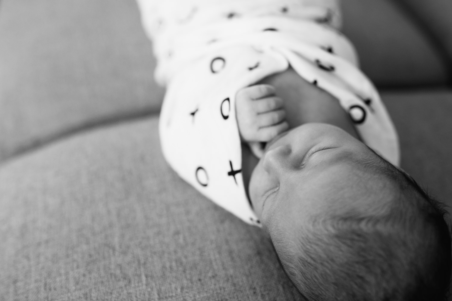 2 week old baby boy with dark hair wrapped in black and white swaddle with arm out, sleeping on grey couch in living room - York Region In-Home Photography