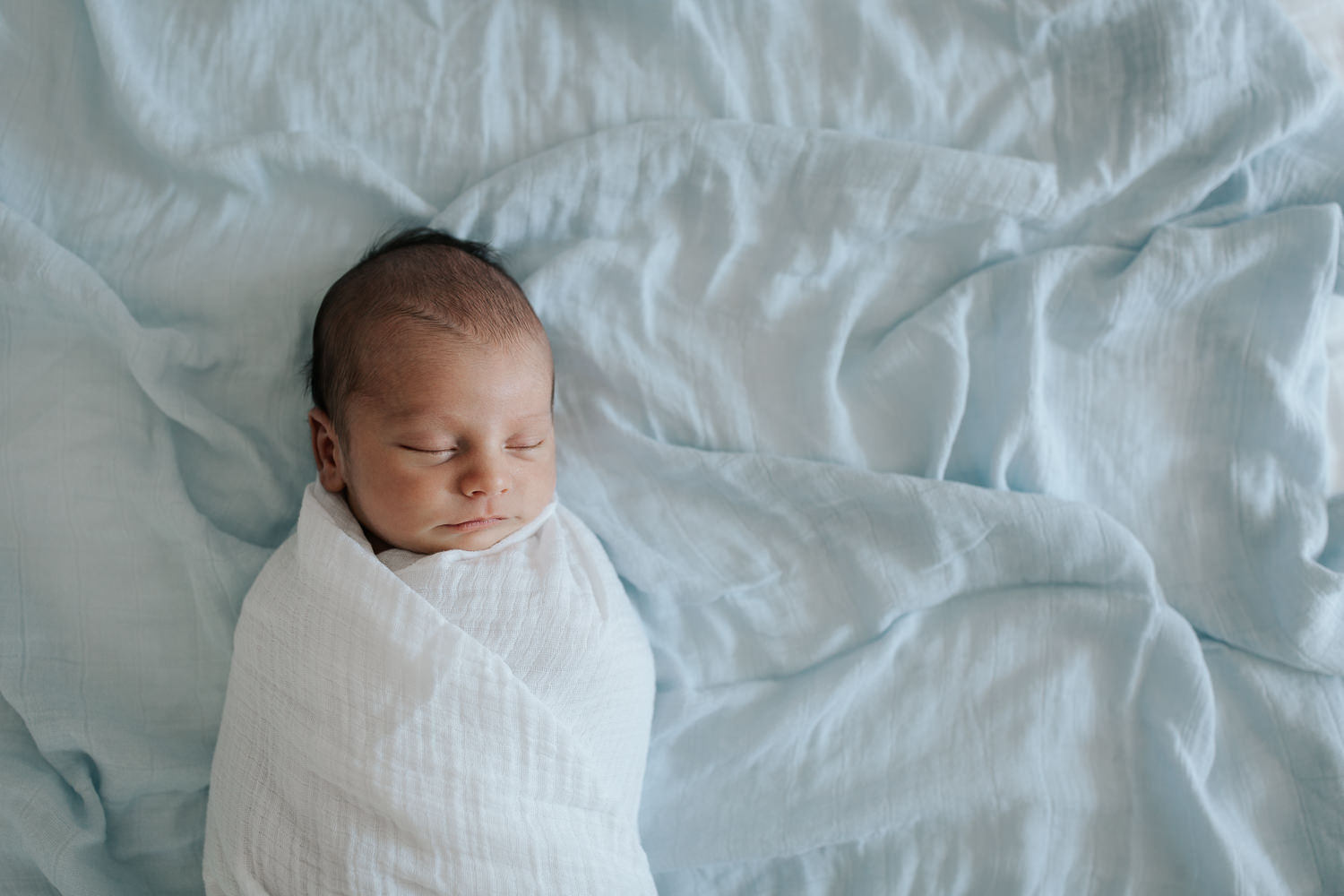 2 week old baby boy with dark hair wrapped in white swaddle lying asleep on bed with blue blanket as background - GTA In-Home Photos