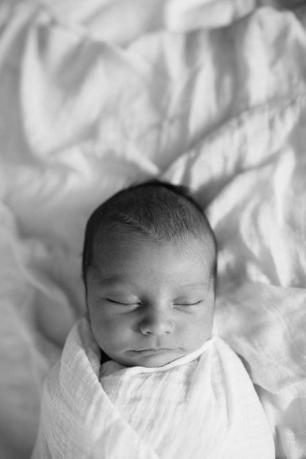 2 week old baby boy with dark hair wrapped in white swaddle lying asleep on bed - Barrie In-Home Photos