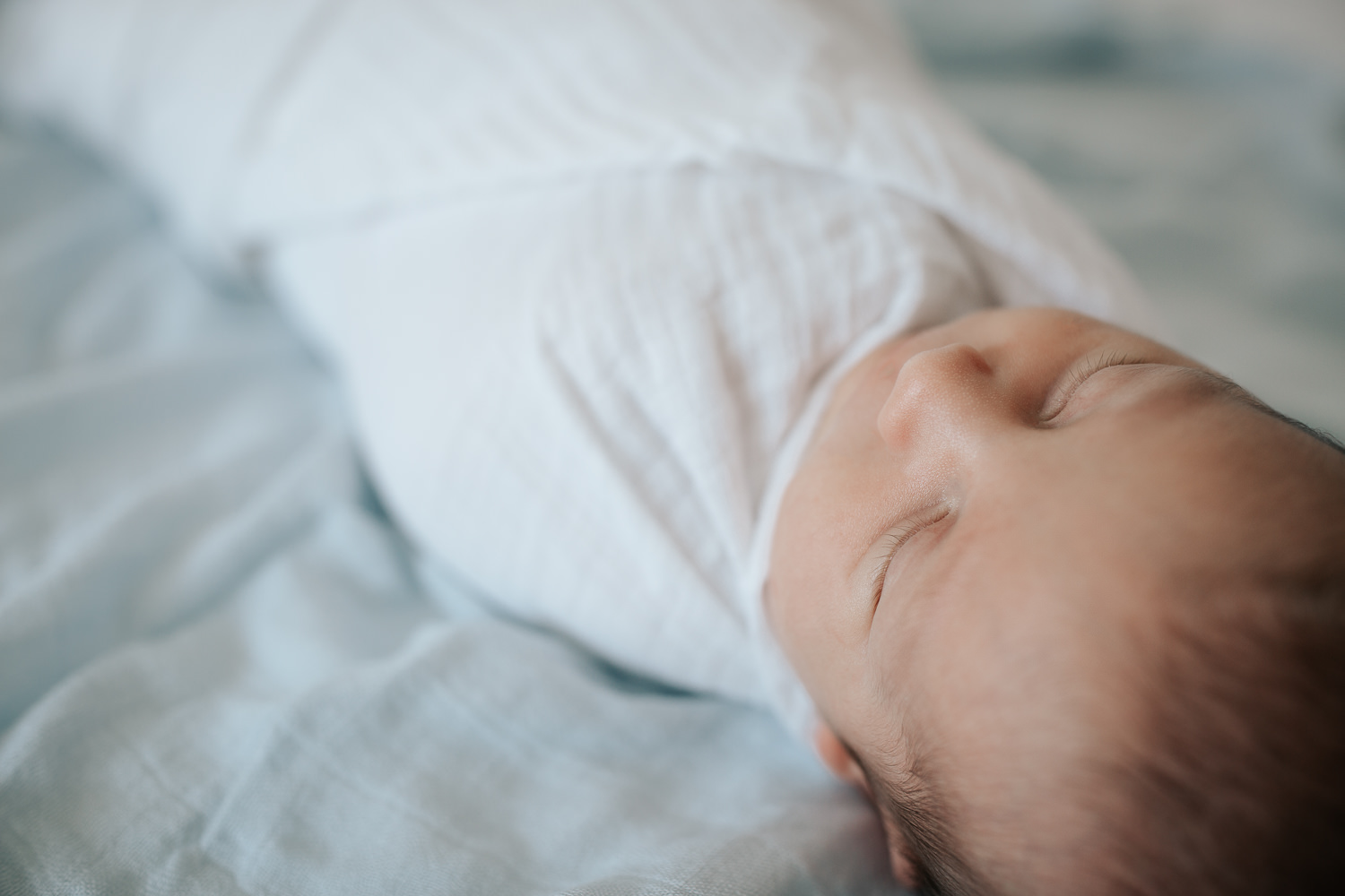 2 week old baby boy with dark hair wrapped in white swaddle lying asleep on bed with blue blanket as background, close up of eyelashes - Stouffville In-Home Photos