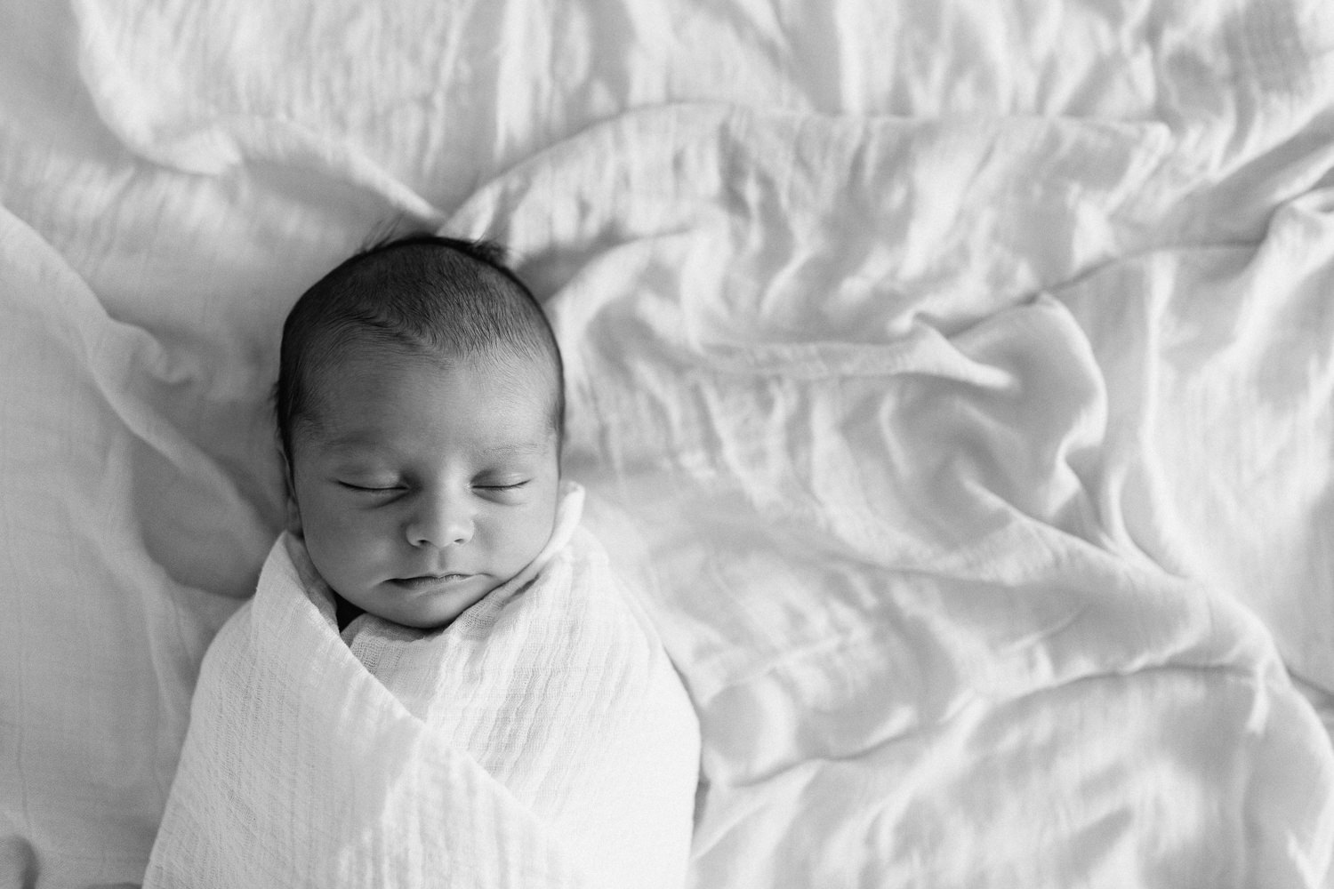 2 week old baby boy with dark hair wrapped in white swaddle lying asleep on bed - York Region In-Home Photos