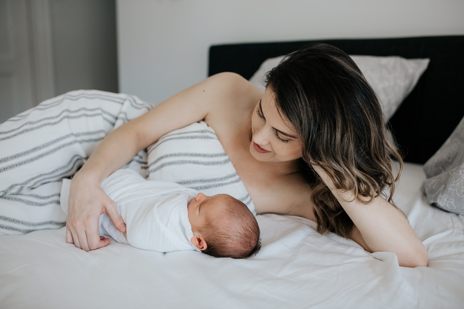 new mother with long dark hair lying on bed next to sleeping 2 week old baby boy wrapped in white swaddle - GTA In-Home Photos